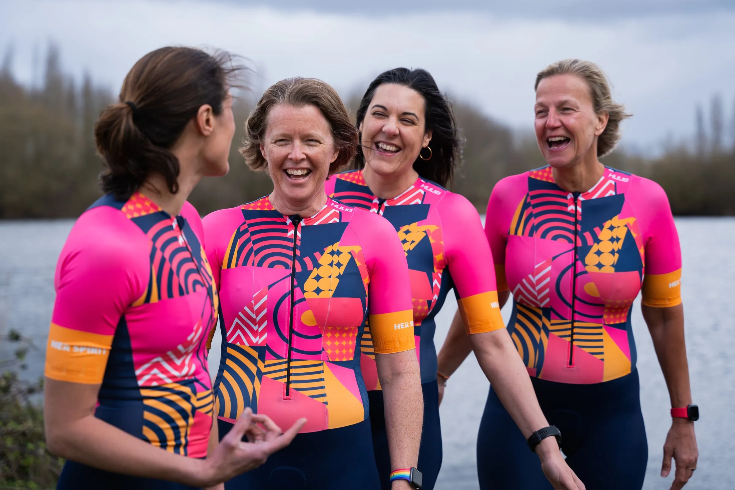 Four laughing women by a lake wearing matching pink, navy and orange HUUB Her Spirit trisuits