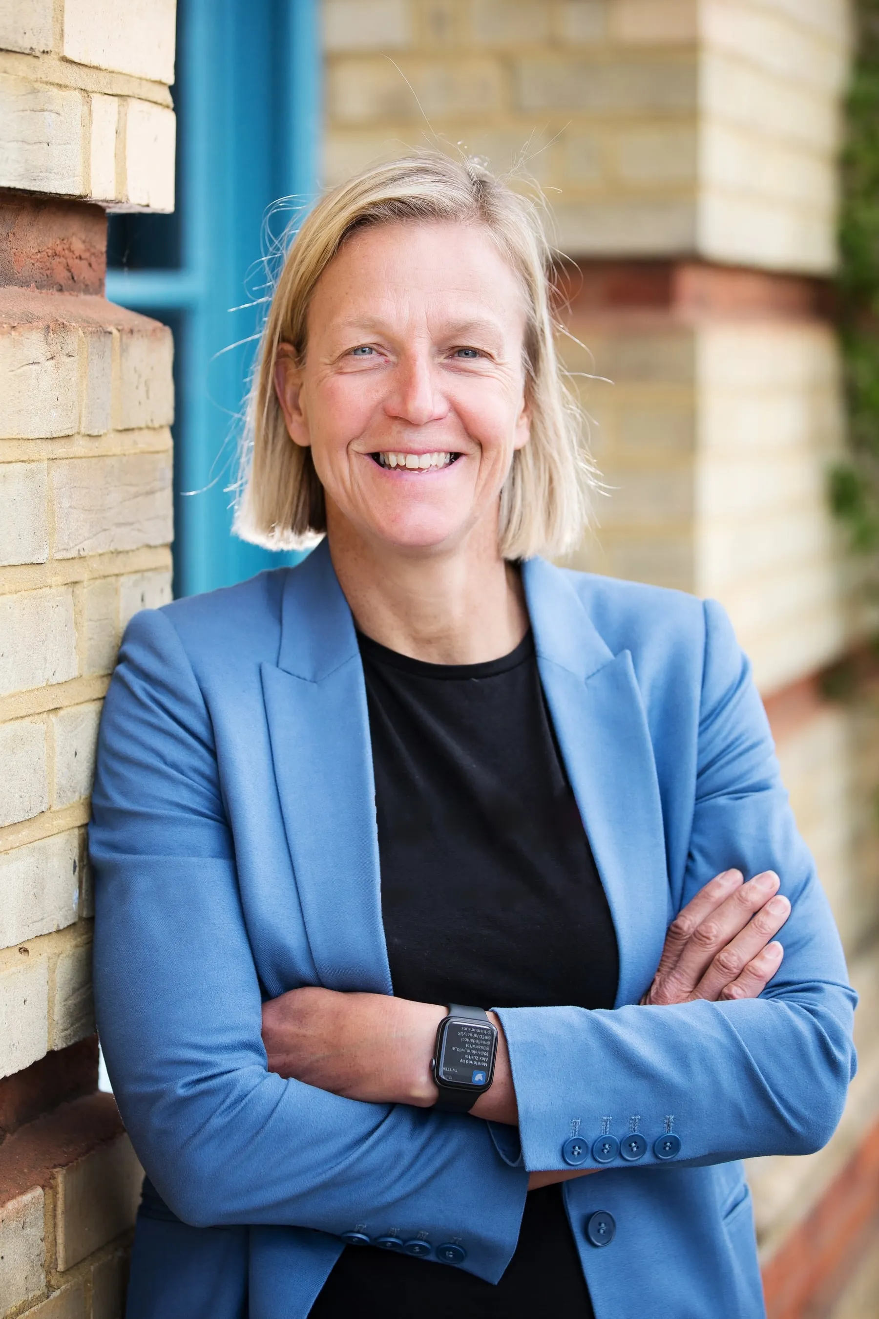 Mel Berry smiling with arms folded, in a blue blazer and a black T-shirt, leaning against a brick wall