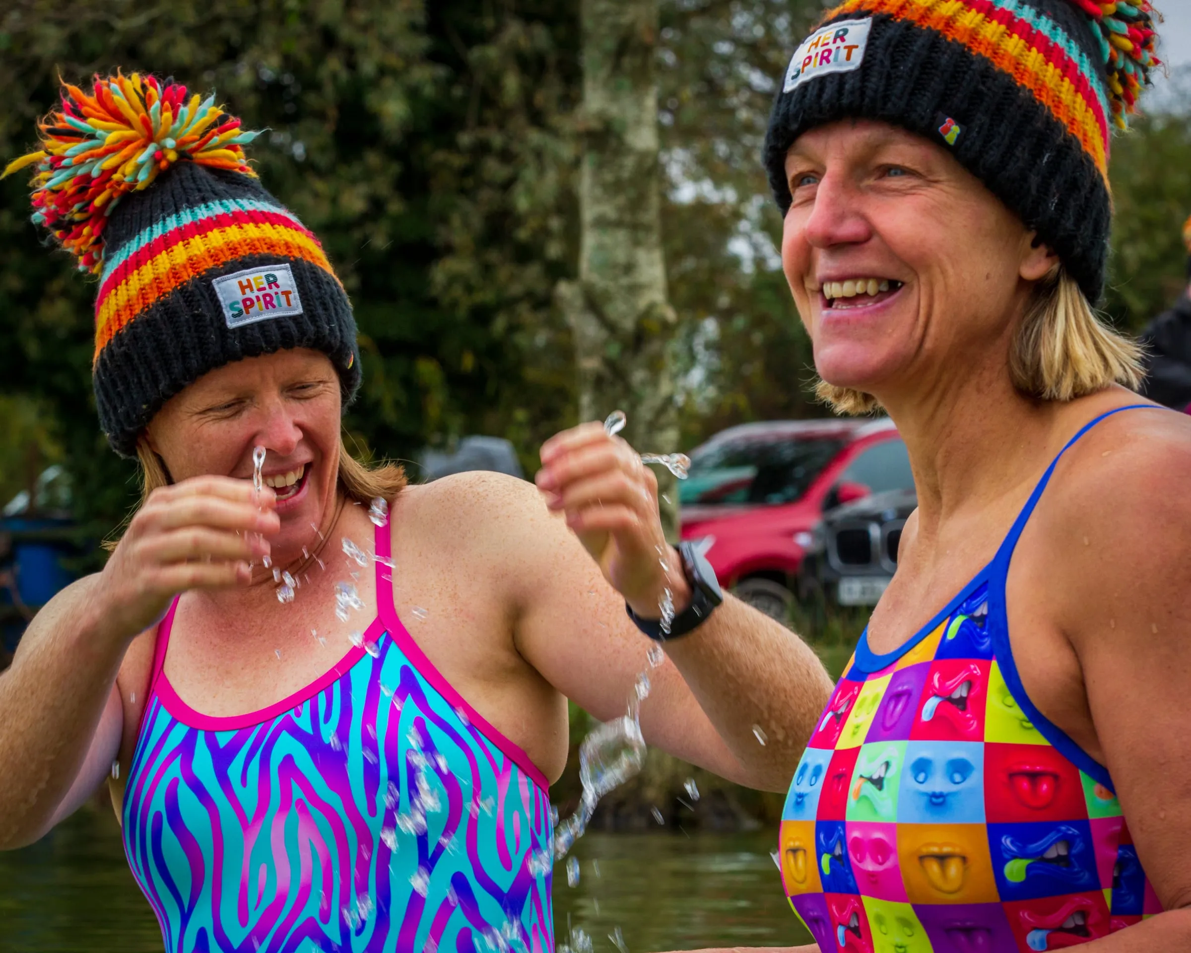 Two women in Her Spirit bobble hats and patterned Zoggs swimsuits laughing and splashing water
