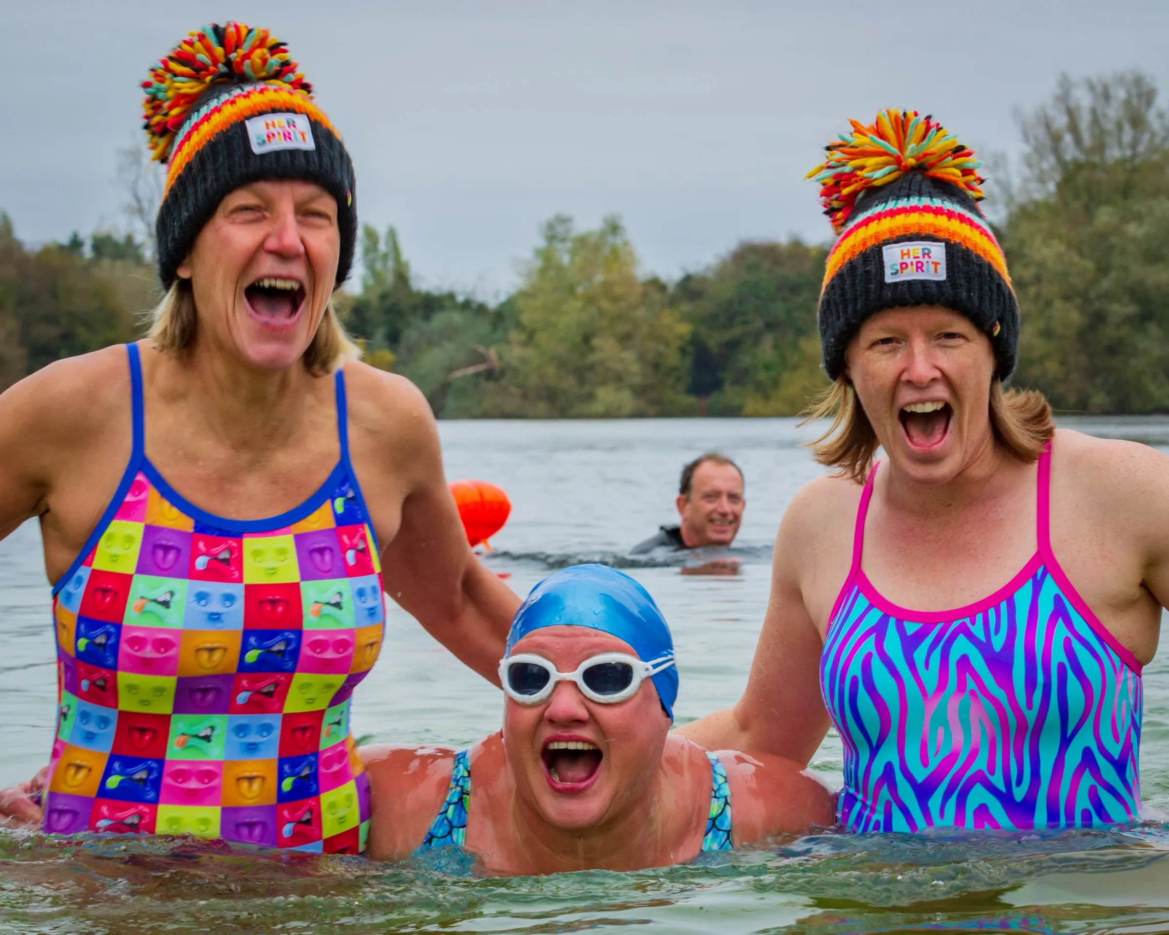 Three swimmers laughing waist-deep in a lake, two in Her Spirit bobble hats and colourful Zoggs swimsuits