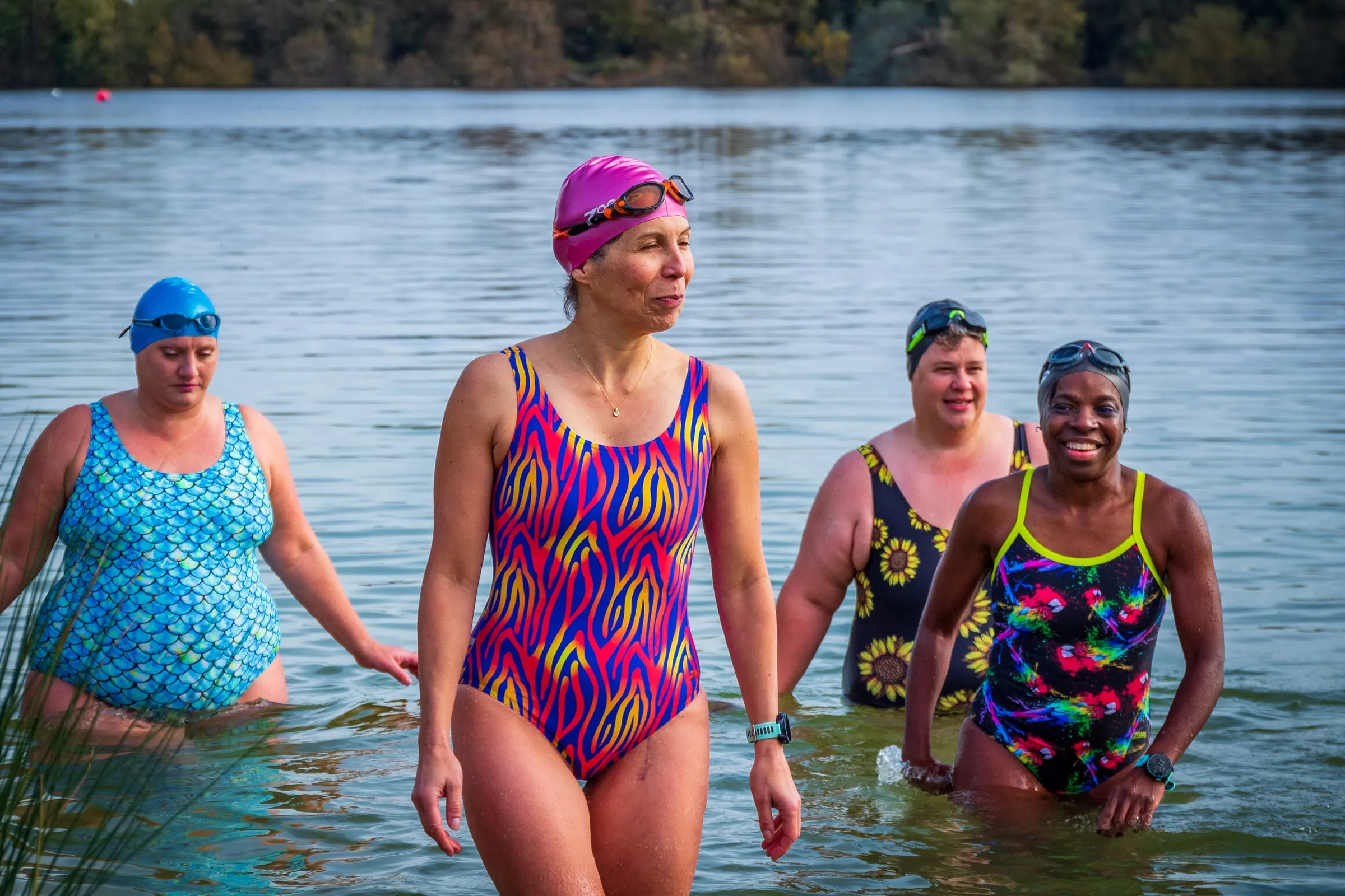 Four women in bright Zoggs swimsuits walking out of a lake after an open water swim, smiling